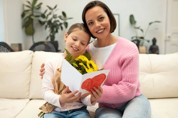 Smiling european millennial woman and little girl with flowers looking at card with heart in living room interior. Gift for mom, birthday greeting, holiday celebration at home, congratulate