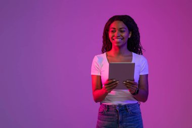 Ellerinde dijital tabletle gülümseyen siyah kadın mor arkaplanının üzerinde neon ışığın içinde duruyor, neşeli Afrikalı genç Amerikalı kadın online alışveriş için modern Gadget kullanıyor ya da serbest çalışıyor.