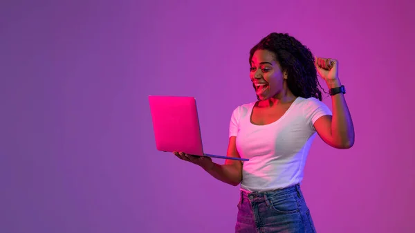 快乐的黑人妇女在霓虹灯下手握手提电脑庆祝成功 快乐的年轻非洲裔美国妇女在电脑上学习 带着兴奋的心情举起拳头 站在紫色的背景下 — 图库照片