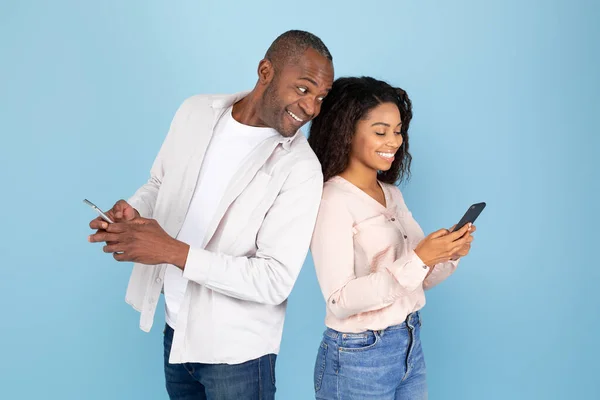 彼らの携帯電話を使って好奇心旺盛な黒人男性は彼女の妻のガジェットと笑顔を見て青いスタジオの背景 — ストック写真