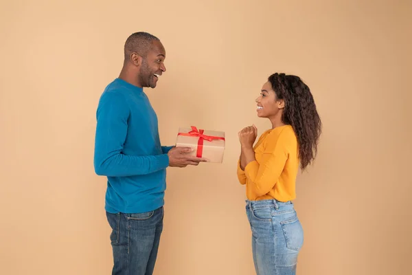兴奋的黑人男人送给妻子礼物 有爱心的中年丈夫送给他兴奋的女人礼物 米色的工作室背景 侧面的观点 生日或周年庆祝活动 — 图库照片