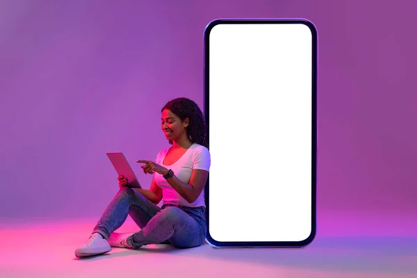 在霓虹灯下坐在大空白智能手机屏风旁的黑人妇女使用数码记事板 在现代家用电器上微笑非洲女性浏览互联网 在紫色背景上张贴照片 拼贴图片 — 图库照片