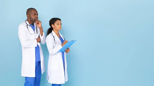 Black male and female doctors thinking, lady holding clipboard looking aside at free space, standing on blue background, panorama. Docs wearing uniform, coat and stethoscopes