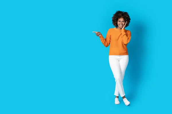 Excited cheerful attractive millennial black lady in casual with bushy hair pointing at copy space and touching her face, posing on blue studio background, showing nice offer, full length
