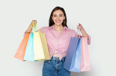 Alışveriş, perakende konsepti. Bir sürü renkli alışveriş çantası taşıyan, gri stüdyo arka planında kameraya gülümseyen ve siyah cuma satışlarının keyfini çıkaran, gündelik kıyafetli mutlu genç bir kadın.