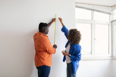 Ev tamiri. Duvarı bant cetvelli ölçen mutlu Afrikalı Amerikalı çift, siyah eşler evlerini yenilerken önlem alıyorlar, kadın kalemle iz bırakıyor, boşluk kopyalıyor.