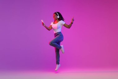 Telsiz kulaklıklı neşeli siyah kadın elinde akıllı telefonuyla zıplıyor, müzik dinleyen heyecanlı Afrikalı Amerikalı kadın ve mor arka planda neon ışığı altında eğleniyor.