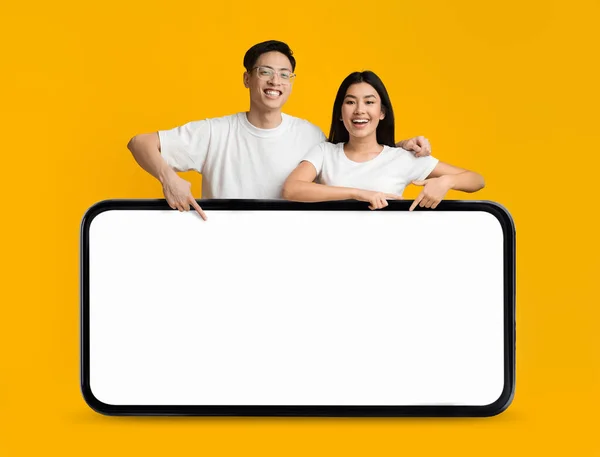 Happy Young Asian Couple Pointing At Huge Smartphone With Blank Screen While Standing Isolated Over Yellow Background, Cheerful Man And Woman Recommending Online Offer Or New App, Collage, Mockup