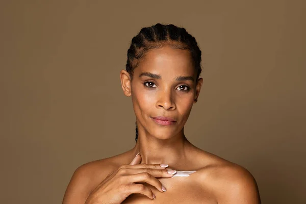African american middle aged woman applying body cream onto her collarbone, posing against brown studio background and looking at camera, copy space