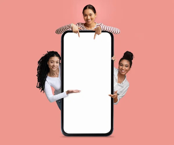 Check This. Three Cheerful Females Pointing At Big Smartphone With Blank Screen, Happy Smiling Women Peeking Out Behind Empty Mobile Phone, Demonstrating Copy Space For Design, Collage, Mockup