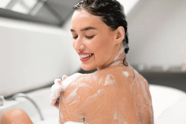 現代的な浴室の浴室の内の浴槽でカメラに戻る肩にシャワージェルを適用泡で体を洗うきれいな女性 ボディケア美容ルーチンコンセプト 選択的フォーカス — ストック写真
