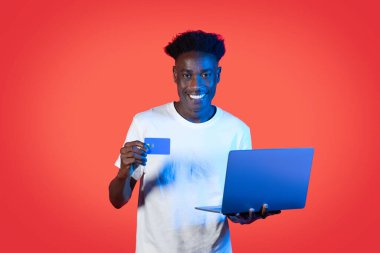 E-ticaret, e-bankacılık. Neşeli, mutlu, beyaz tişörtlü, elinde modern dizüstü bilgisayar ve banka kartı olan genç, neon ışıklı kırmızı stüdyo arkaplanı üzerinden mal ve hizmetler için ödeme yapan, neşeli, mutlu bir siyah adam.