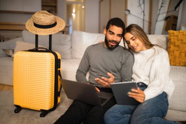 Gülümseyen, çok kültürlü genç çift ellerinde aletler, bavul ve şapkayla yerde oturuyor ve oturma odasında tatil için bir ülke seçiyorlar. Seyahat planı, hazırlık uygulaması, bilet parası.