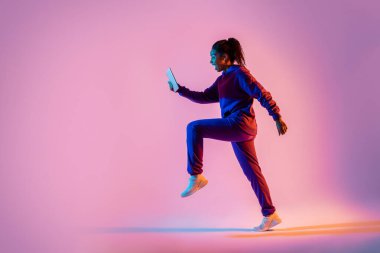 Genç, sportif, atlayan, koşan ve cep telefonunu pembe ışıklı arka plan, yan görüş, kopyalama alanı üzerinden kullanan kadının vücut boyu.