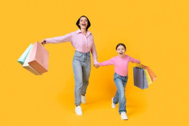 Aile alışverişi. Mutlu anne ve küçük kızı, sarı stüdyo geçmişinde parlak alışveriş torbalarıyla geziyor, neşeli anne ve kız çocuğu mevsimlik satışlardan ve indirimlerden keyif alıyor,