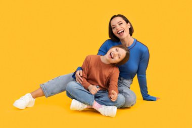 Anne ve kızı. Sarı stüdyo arka planında izole edilmiş bir şekilde otururken mutlu bir anne ve kızın portresi ve annesi gülüyor ve eğleniyor.