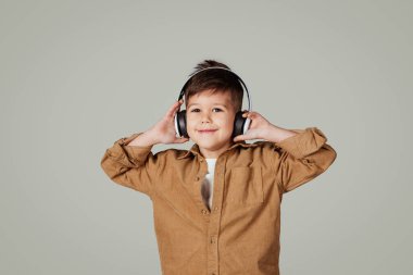 Kablosuz kulaklıklı gülümseyen küçük Avrupalı çocuk müzik dinliyor, gri stüdyo arka planında izole edilmiş boş zamanın tadını çıkarıyor. Okul çocuğu ses uygulaması, öğrenim ve eğitimle iyi eğlenceler.