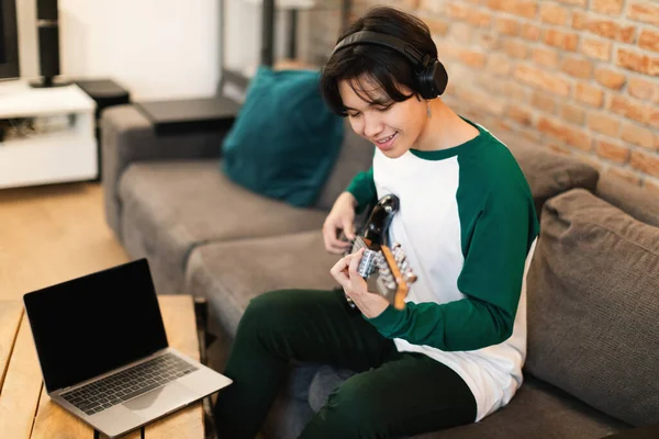 陽気な日本のティーン男の子は週末に自宅でソファに座って楽器をオンラインでプレイするためにノートパソコンの学習の近くに電気ギターでコードを再生します 音楽趣味の概念 選択的フォーカス — ストック写真