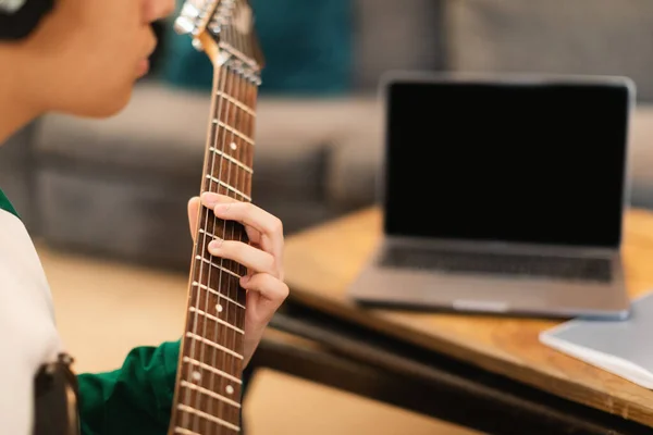 家庭でノートパソコンと机に座ってエレクトリックギターを演奏イヤフォンを身に着けているアジアの十代の男 週末に新しいコードを学ぶ才能のある少年 トリミングされたショット 選択的なフォーカス — ストック写真