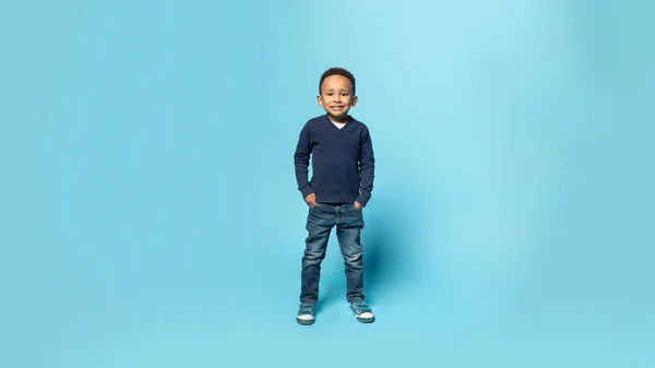 快乐的童年全长拍摄可爱的非洲裔美国小男孩手插在口袋里 在蓝色的摄影棚背景上摆出姿势 对着相机微笑 全景尽收眼底 — 图库照片