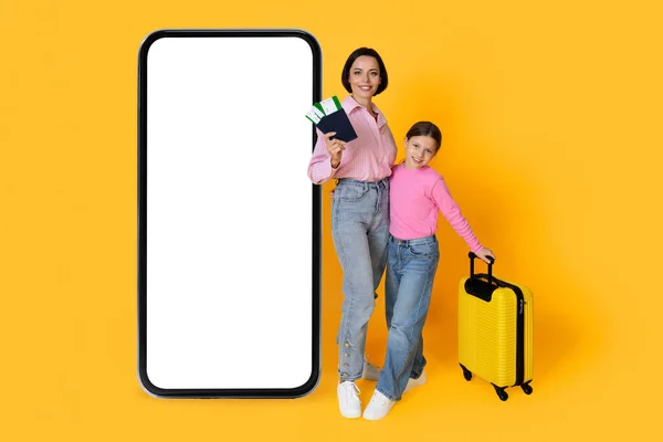 白い画面で大きな空白のスマートフォンの近くに立ってスーツケースとチケットを持つ幸せな母と娘 笑顔家族旅行予約のための新しいモバイルアプリ 創造的なコラージュ モックアップ — ストック写真