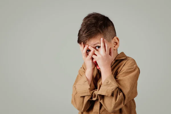 Frightened little european boy covers his face with hands and afraid isolated on gray studio background. Schoolboy emotions, fear, horror and stress, study and education at school