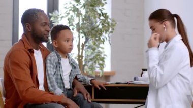 Hastalık muayenesinden sonra. Dost canlısı bayan doktor klinikte nefes alan küçük çocuğu dinliyor, akciğerlerini kontrol ediyor, Afrikalı Amerikalı çocuk babasıyla klinikte oturuyor.