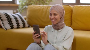 İnternet iletişimi. Genç mutlu ortadoğulu bayan geleneksel türban ağı takıyor evde akıllı telefondan sörf yapıyor, sosyal medyayı evde kaydırıyor, çekim takip ediyor, yavaş çekim, boş alan