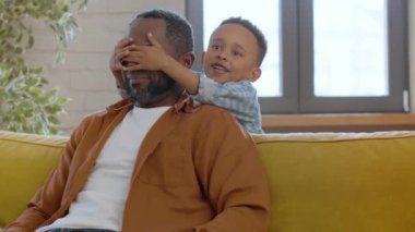 Aile eğlencesi. Neşeli Afrikalı Amerikalı adam evdeki kanepede dinleniyor, tatlı küçük oğlu geliyor ve gözlerini kapıyor, hafta sonları birlikte gülüyor, çekim takip ediyor, boşluk