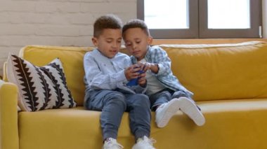 Çocuklar ve modern aletler. İki Afrikalı Amerikalı erkek kardeş akıllı telefondan video oyunu oynuyorlar. Cihazı paylaşıyorlar. Evdeki koltukta oturuyorlar.