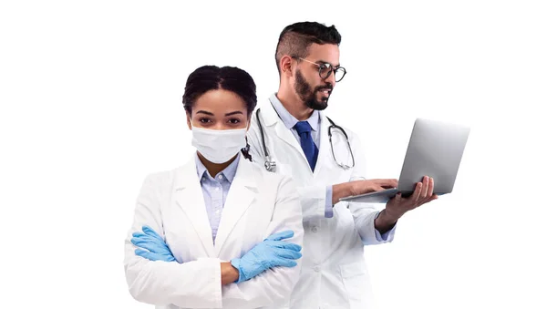 阿拉伯医务工作者使用手提电脑 而黑人女医生则用照相机和胶水来观察 这是由两名身穿制服 与白人背景隔离的多族裔医生组成的综合保健形象 — 图库照片