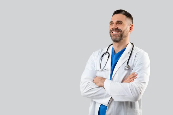 灰色のスタジオの背景に折り畳まれた腕で制服姿の男性医師を自信を持って 自由空間を見て 首の周りに聴診器を持つ医師笑顔 — ストック写真