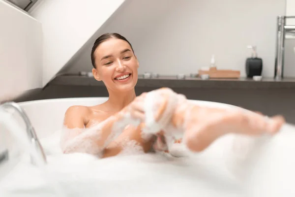 お風呂に入る幸せな女性お風呂ブラシマッサージで脚をこすり 自宅のモダンなバスルームで泡でいっぱいのバスタブに座って体を洗う ボディケアの概念 選択的フォーカス — ストック写真