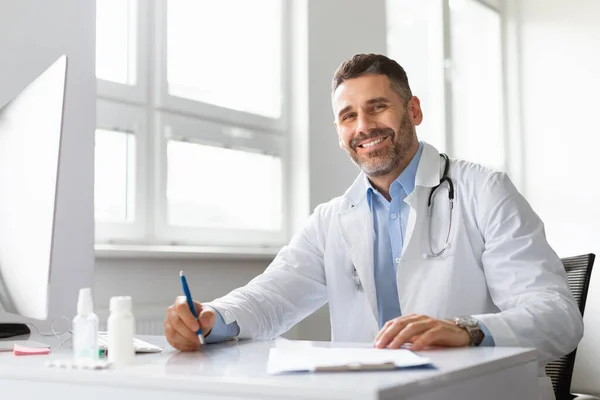 病院の机に座ってカメラで笑顔し コピースペースで白い医療制服を着た幸せな中年男性医師 診療所の医学雑誌で博士号を取得 医学の概念 — ストック写真