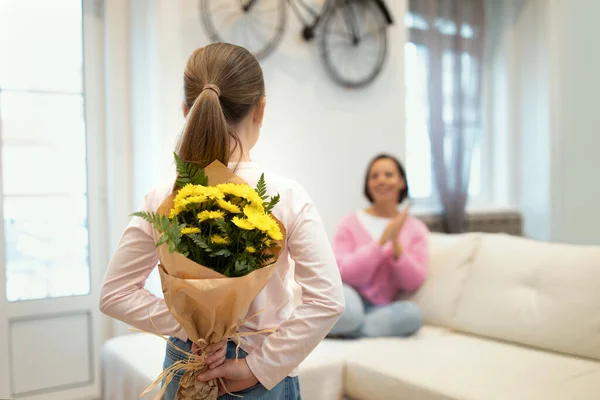 Happy european adult woman looking at little girl with flowers in white living room interior, blurred, free space. Gift for mom, birthday greeting, holiday celebration at home