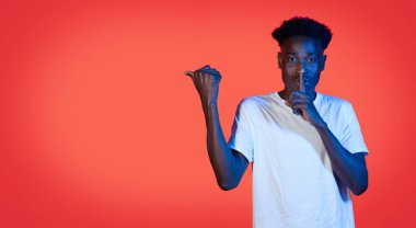 Beyaz tişörtlü yakışıklı siyahi adam parmağını dudaklarına uzatıp sessizliğini koruyor ve kırmızı stüdyo arkaplanındaki kırmızı ışıkta reklam için fotokopi alanını işaret ediyor.