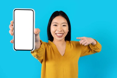 Gülümseyen güzel Asyalı kadın elinde beyaz ekranlı modern bir telefon tutuyor. Güzel bir mobil uygulama ya da online teklif sunuyor. Mavi stüdyo arka planında izole edilmiş.