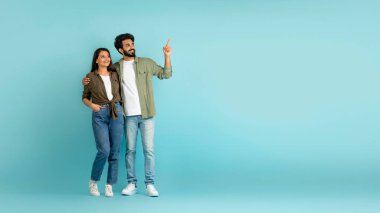 Orta Doğulu sıradan bir erkek ve Hintli kadını kucaklayan ve mavi stüdyo arka planında reklam yapmak için boş alana bakan neşeli, güzel, çok ırklı bir çift.