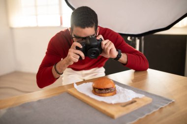 Yemek fotoğrafçısı yakışıklı, orta doğulu günlük kıyafetler giyen ve gözlüklü genç bir adam fotoğraf stüdyosunda lezzetli etli hamburgerin fotoğraflarını çekiyor, video kamera kullanıyor.