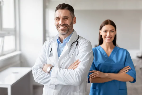 Happy male doctor and female nurse colleagues, wearing coats, standing with folded arms, posing and smiling at camera at modern light medical office