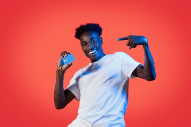 Neşeli, yakışıklı, milenyum siyahi adam mavi plastik banka kartına bakıyor ve gülümsüyor. Temassız ödeme öneriyor. Kolay hızlı bankacılık, kırmızı stüdyo arka planı, neon ışığı, kopyalama alanı.