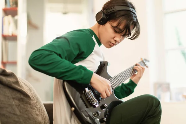 Japanese Teen Guy Wearing Headphones Learning Electric Guitar Sitting On Sofa In His Room At Home. Musician Boy Playing Musical Instrument Writing New Song On Weekend. Selective Focus