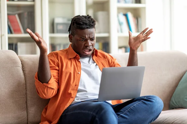 Furious young black chubby man entrepreneur reading emails on laptop at home, gesturing. African american guy sitting on beige couch in living room, having troubles, copy space