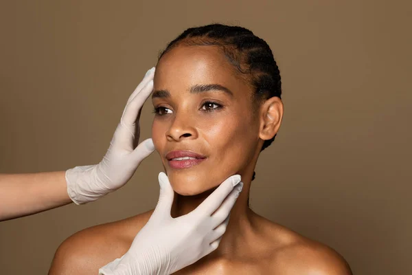 中年妇女整容 医生手拿着医用手套 手持黑人女病人的头 在手术前检查皮肤 棕色工作室背景 — 图库照片