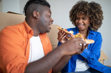 Neşeli siyahi çift pizza yiyor ve taşınma gününde eğleniyor, romantik Afrikalı Afrikalı Amerikalı eşler, karton kutular arasında dinlenirken Ait oldukları eve taşınıyor, kadınları doyuran koca, yakın plan