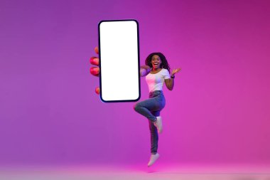 Harika bir teklif. Elinde büyük, boş, akıllı bir telefonla atlayan heyecanlı siyah kadın, Mor Arkaplan üzerinde neon ışığı pozu verirken beyaz ekranlı cep telefonu gösteren sürpriz Afrikalı kadın,