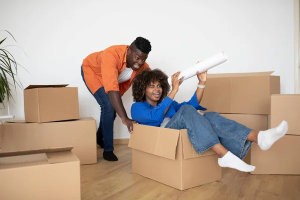 Cheerful Black Couple Having Fun While Relocating Home, Man Pushing Cardboard Box With His Happy Wife, Happy African American Spouses Fooling Together While Unpacking Things On Moving Day