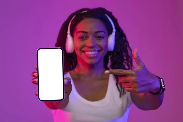 Online Ad. Happy Black Female Pointing At Blank White Smartphone Screen, Smiling African American Woman In Headphones Recommending Music App, Standing In Neon Light Over Purple Background, Mockup