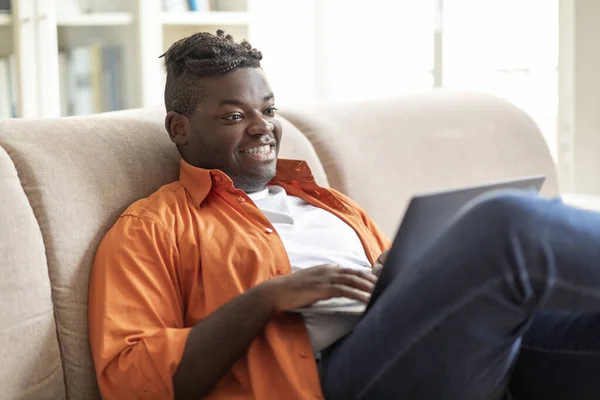 Cheerful happy relaxed young african american guy in casual sitting on couch at home, using modern pc laptop, typing on computer keyboard, websurfing, chatting, looking for job online, copy space