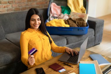 Kredi kartı ve dizüstü bilgisayar kullanan, yurt dışına seyahat rezervasyonu yapan ya da otel rezervasyonu yapan, online seyahat acentesinin internet sitesinde gezinen, paketlenmemiş valizin yanında oturan İspanyol bir kadın.